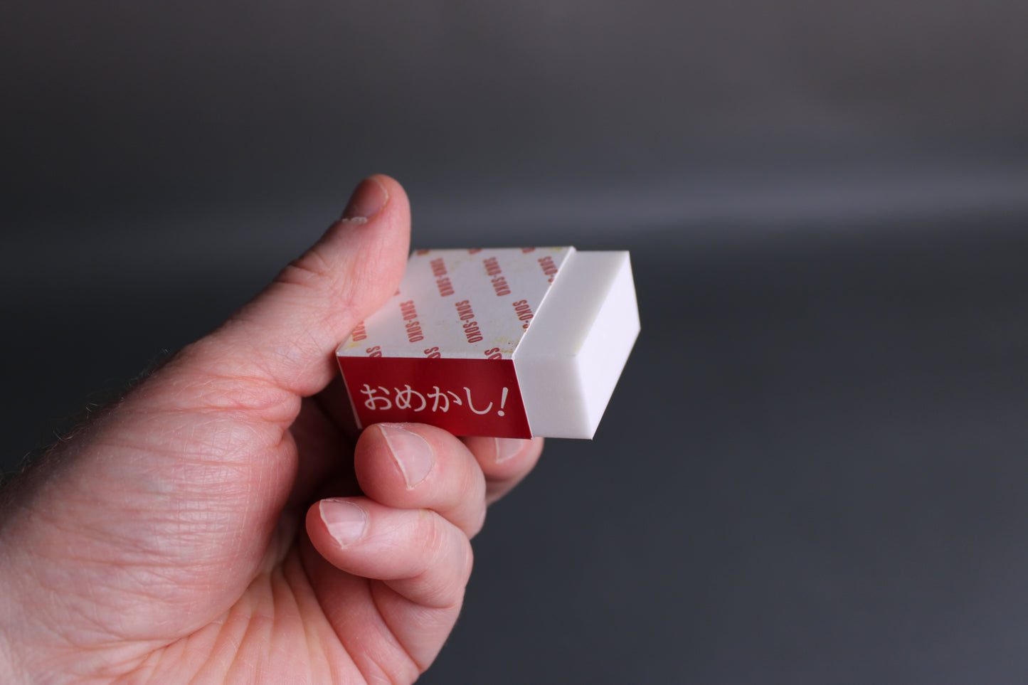hand holding white rectangular block rust eraser with paper wrapper cover with hiragana omekashi grey backdrop