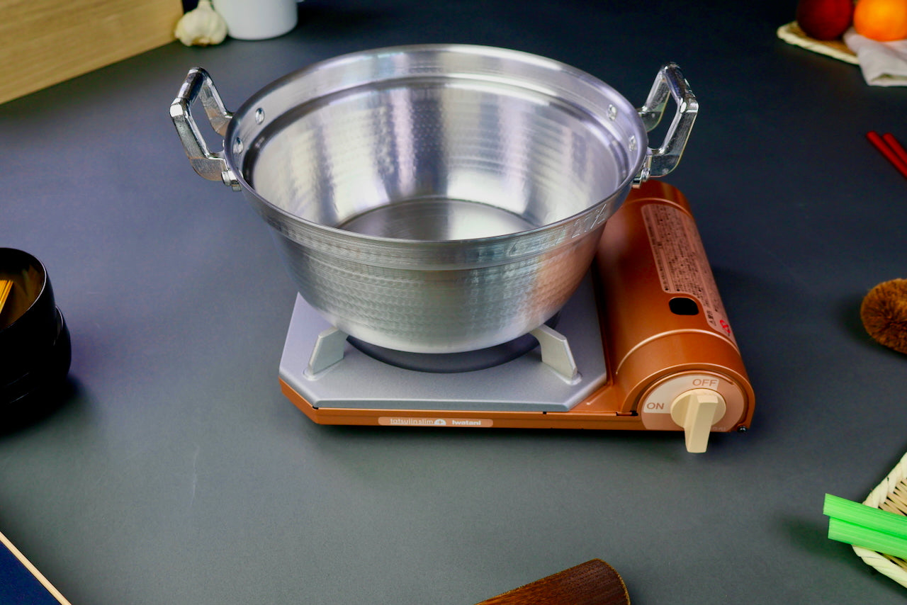 two handled stepped aluminum silvery pot with shiny hammered surface sits atop portable silver tan butane stove surrounding kitchenware dark grey backdrop