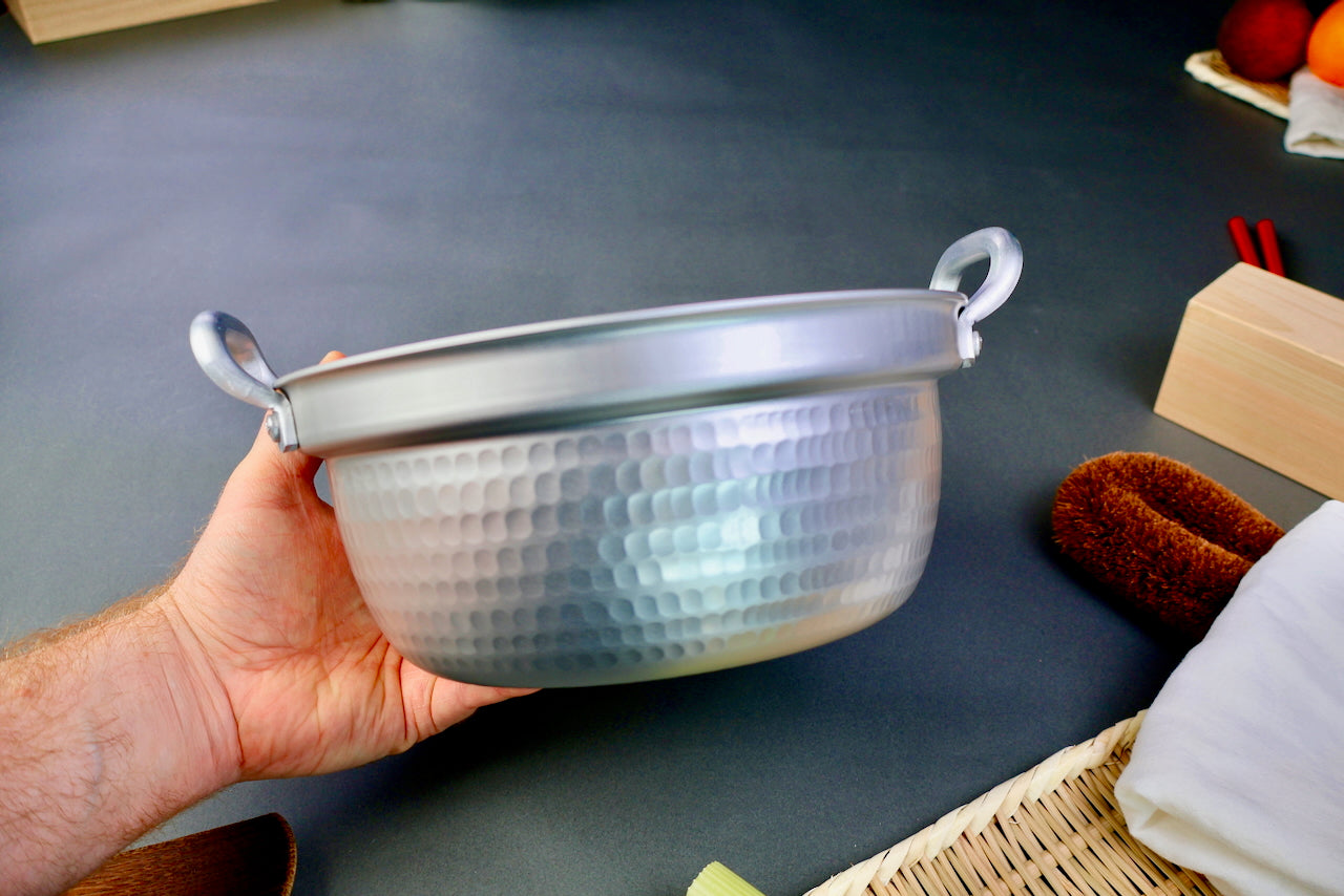 hand holding stepped aluminum silvery pot with shiny hammered surface surrounded by japanese kitchenware on a dark grey backdrop