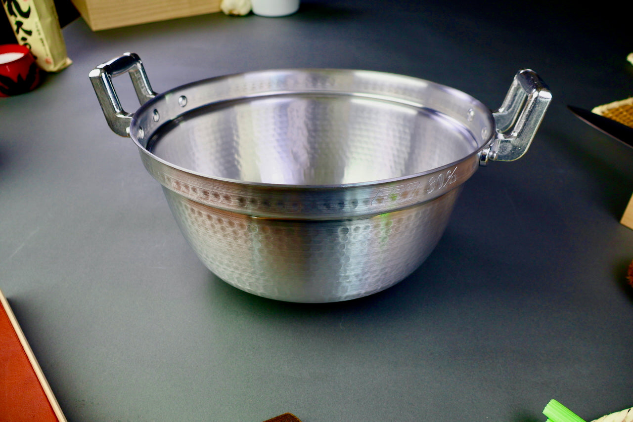 two handled stepped aluminum silvery pot with shiny hammered surface surrounded by japanese kitchenware on a dark grey backdrop