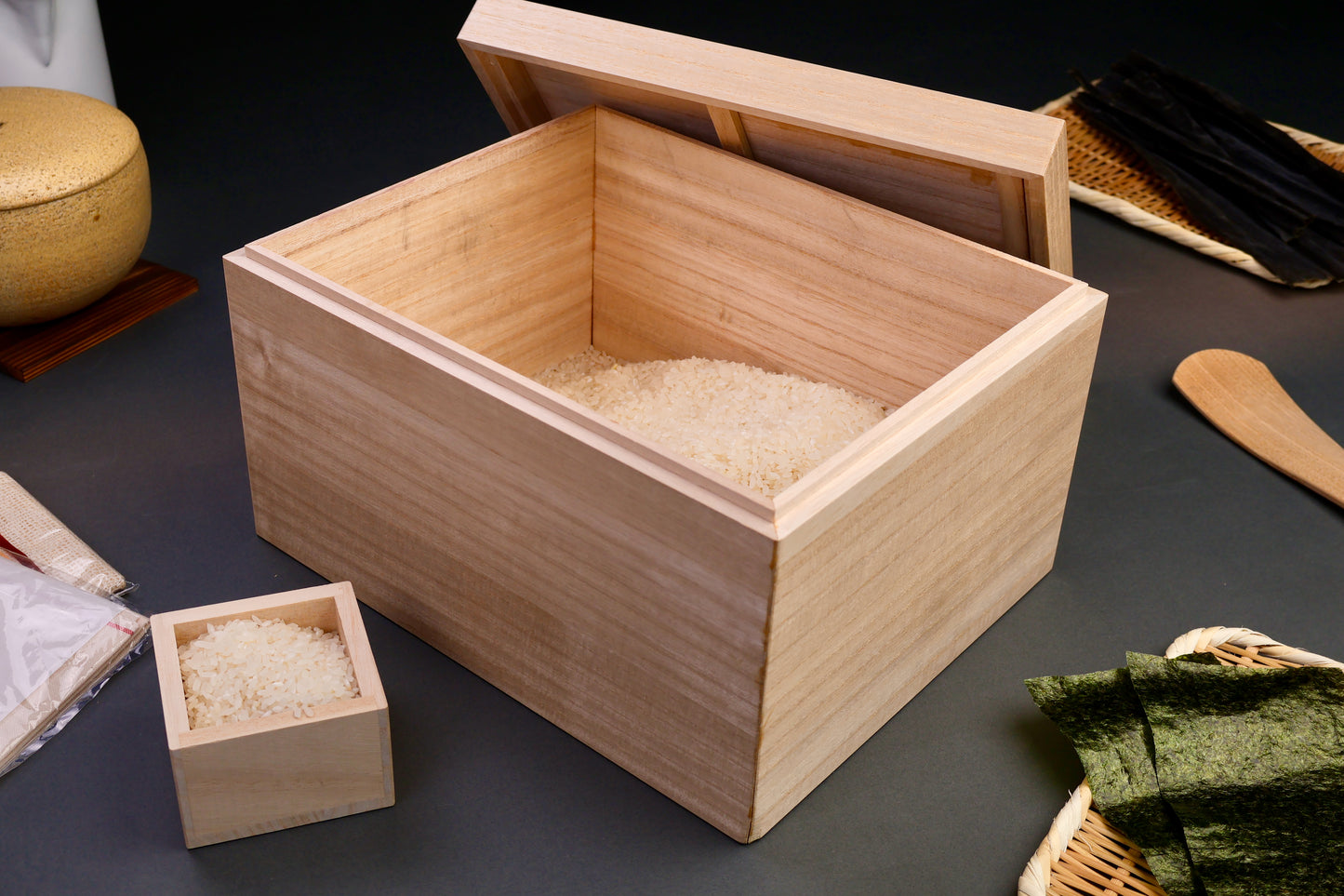 tall rectangular paulownia wood rice box sliding door on top removed with rice scoop surrounded by japanese kitchenware dark grey background