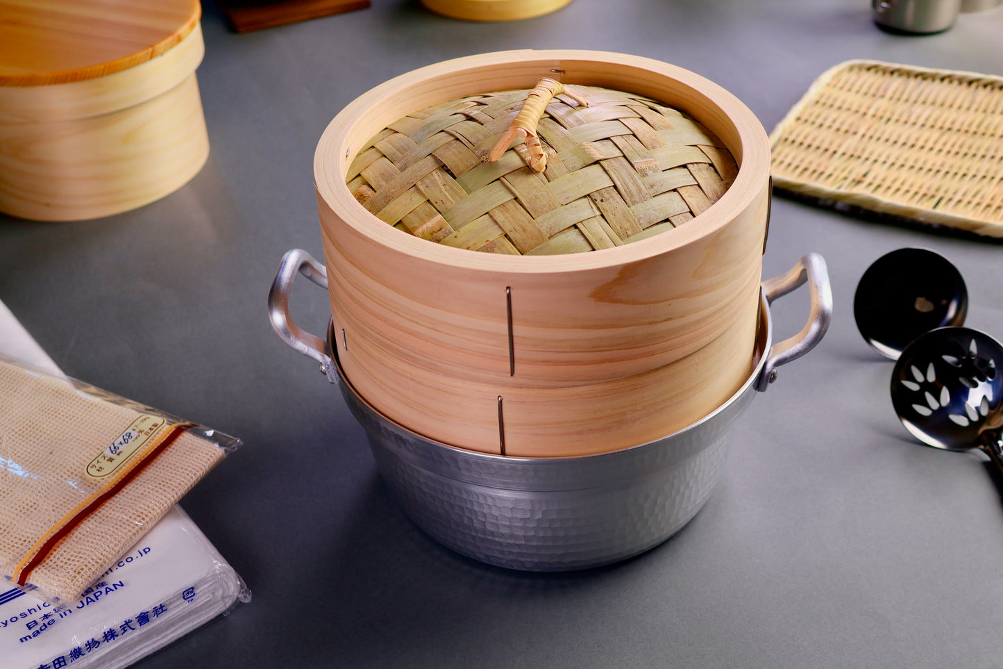 frontal shot of hinoki wood steamer showing elongated wood grain pattern sits atop aluminum two handled stepped pot surrounded by japanese kitchenware