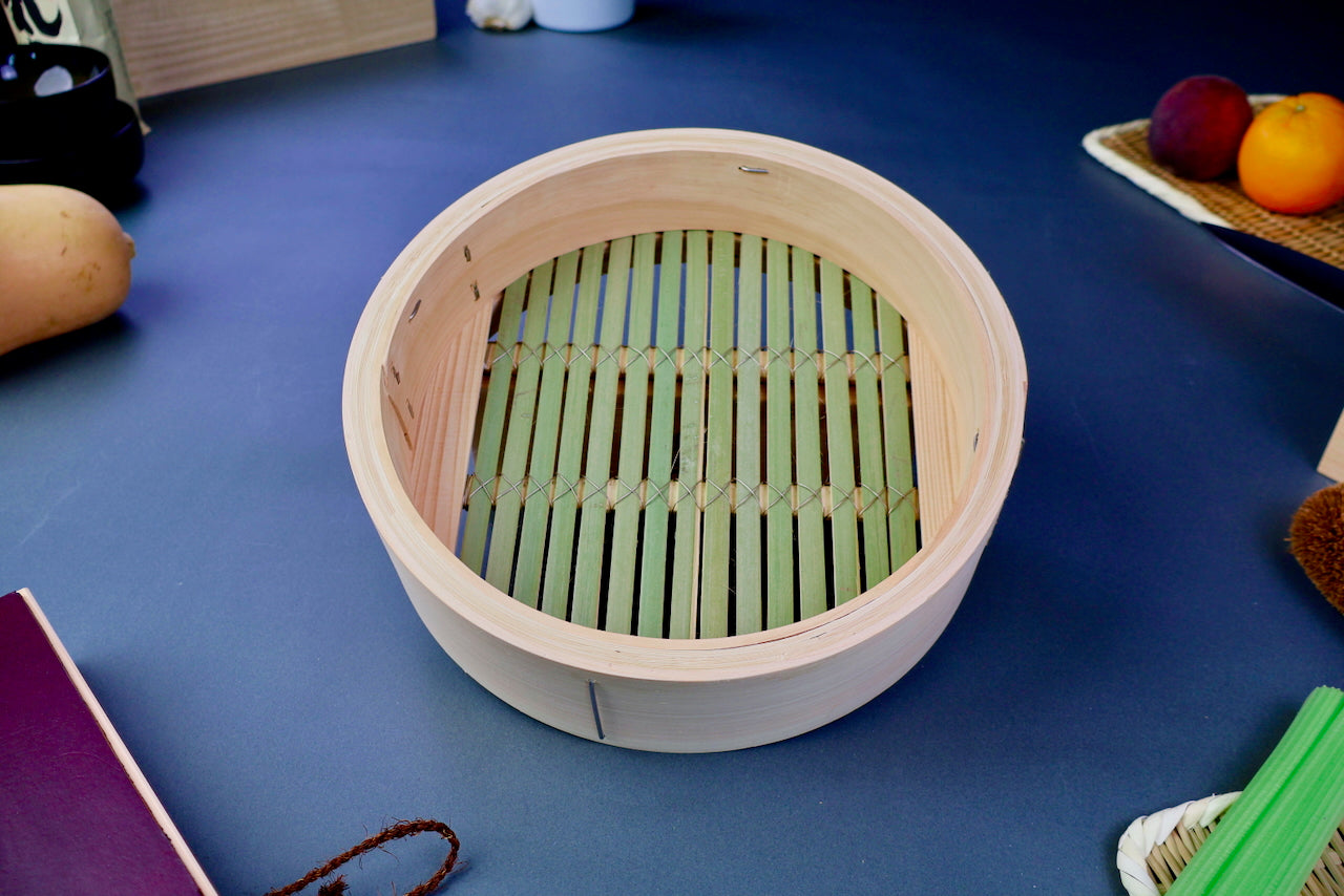 round thick hinoki wood bamboo steamer base with thick greenish slats interior surrounded by japanese kitchenware on blue gray surface