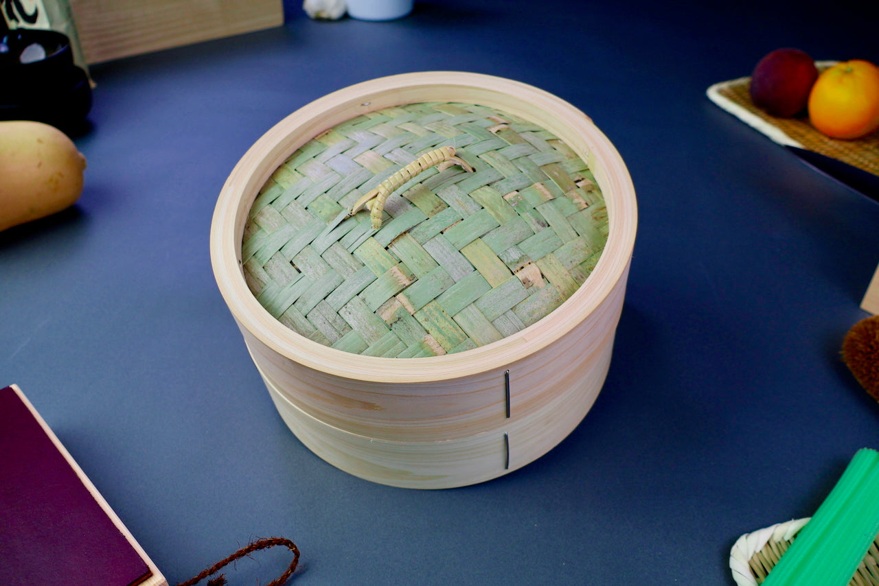 thick wood round bamboo steamer set woven top with loop natural wood tone with printed hiragana surrounded by japanese kitchenware in blue gray background