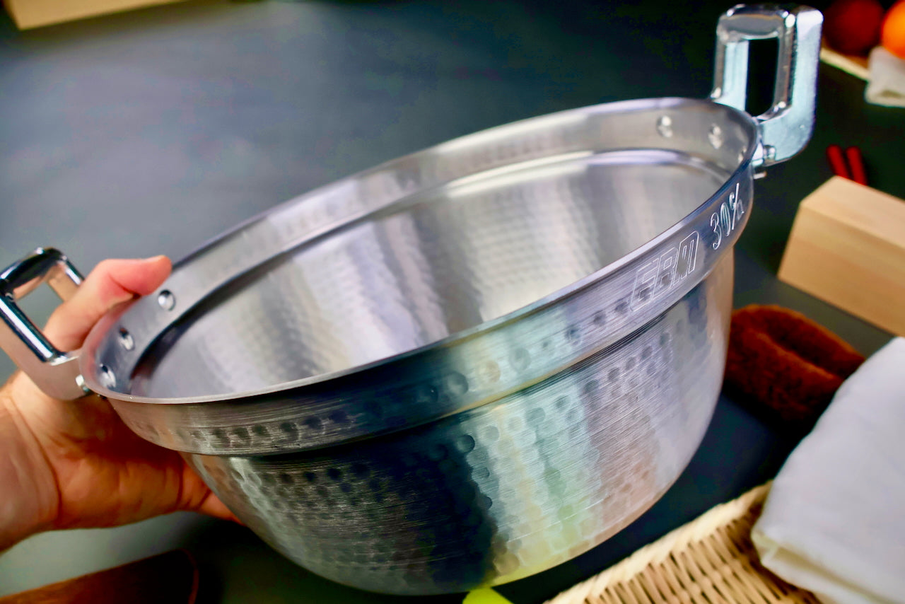 hand holding a two handled stepped aluminum silvery pot with shiny hammered surface surrounded by japanese kitchenware on a dark grey backdrop