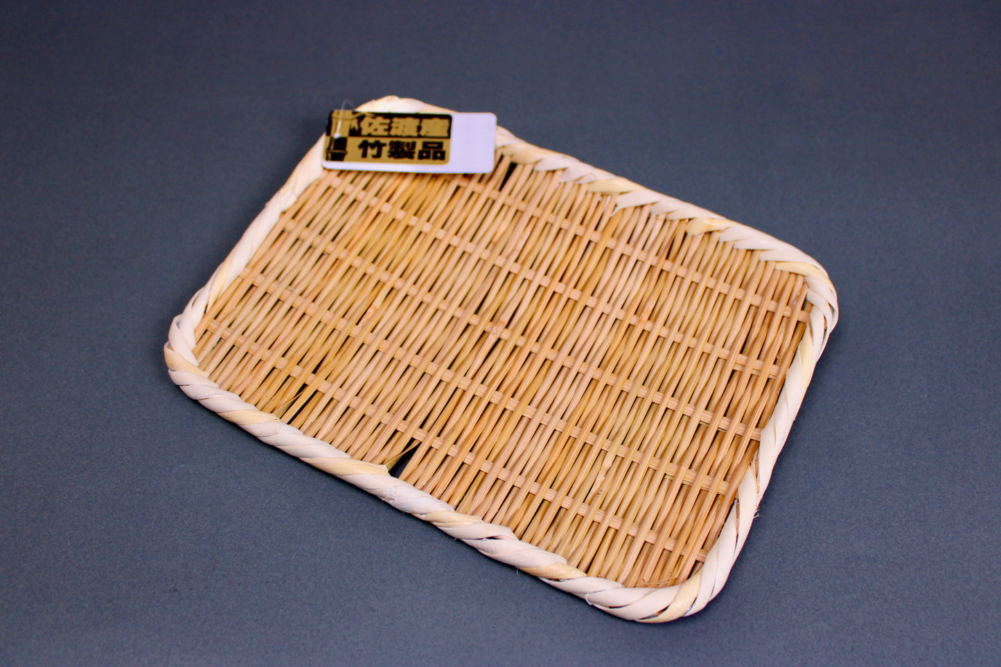 woven bamboo strainer rectangular tan brownish in color with product tag showing contrasting gold black kanji characters in grey blue backdrop