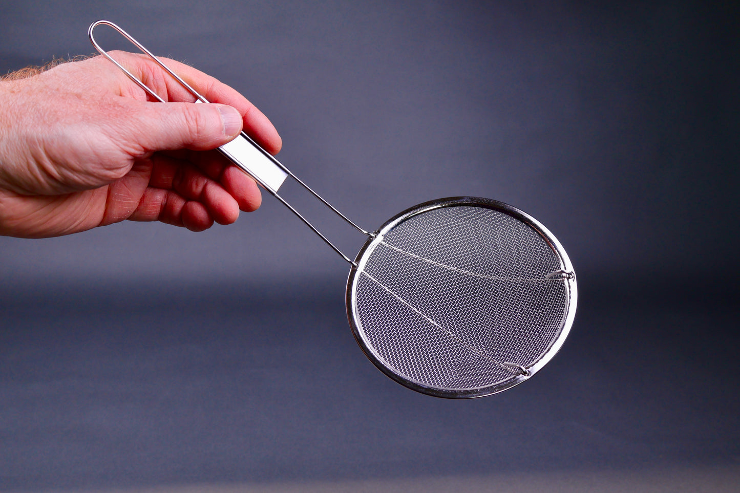Metal coarse skimmer with long thin handle held by a hand against a dark background
