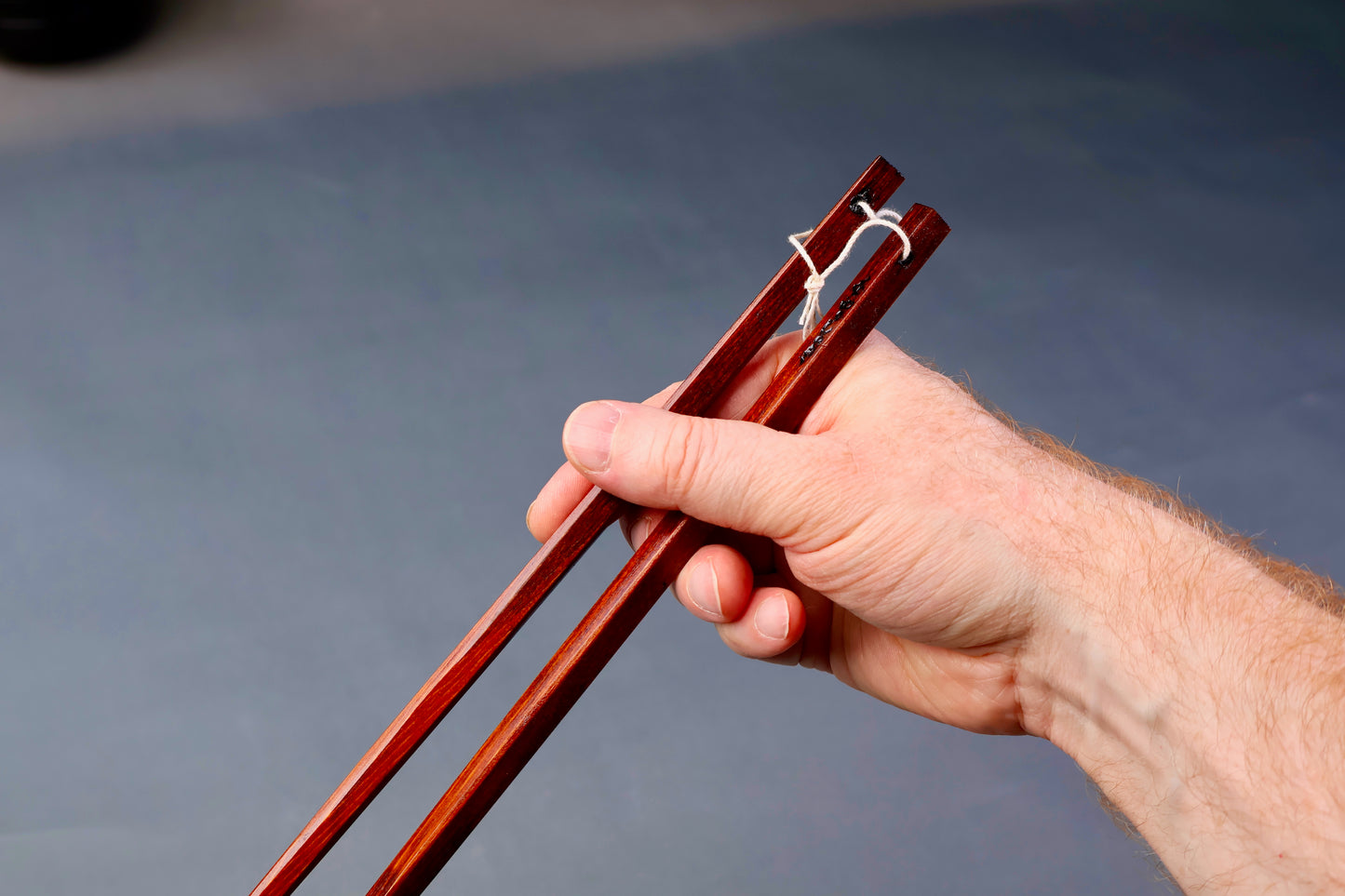 hand holding long square brownish tan hinoki wood cooking chopsticks urushi lacquered connected with white twine surrounded bluish grey background