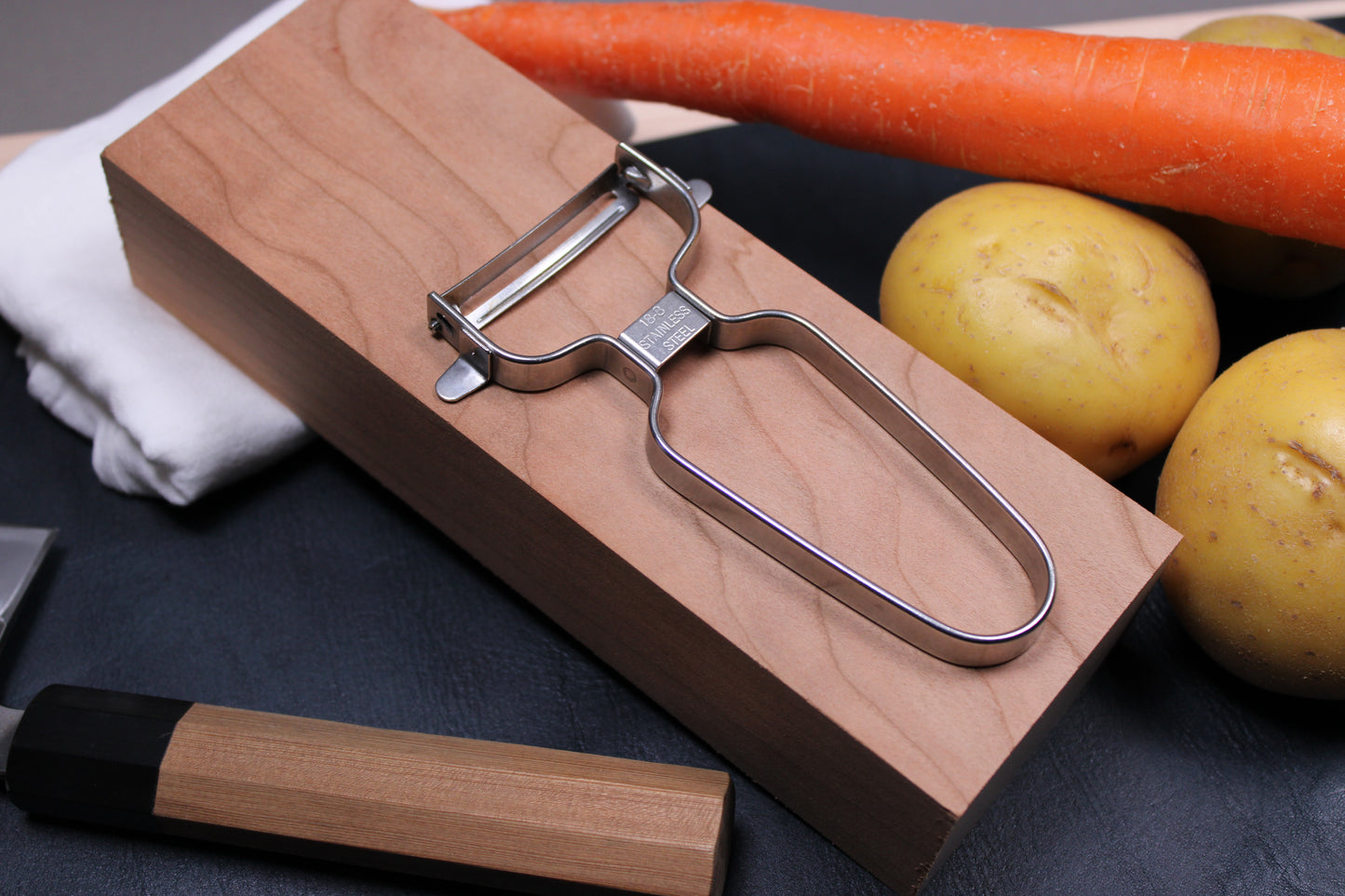 stainless steel vegetable peeler by shimomura atop cherrywood block beside cedar handled nakiri knife yellow potatoes carrots gray background