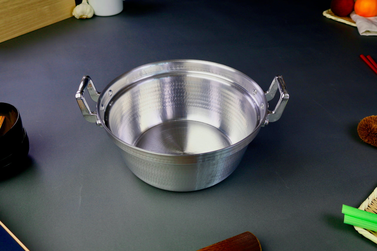 two handled stepped aluminum silvery pot with shiny hammered surface surrounded by japanese kitchenware on a dark grey backdrop
