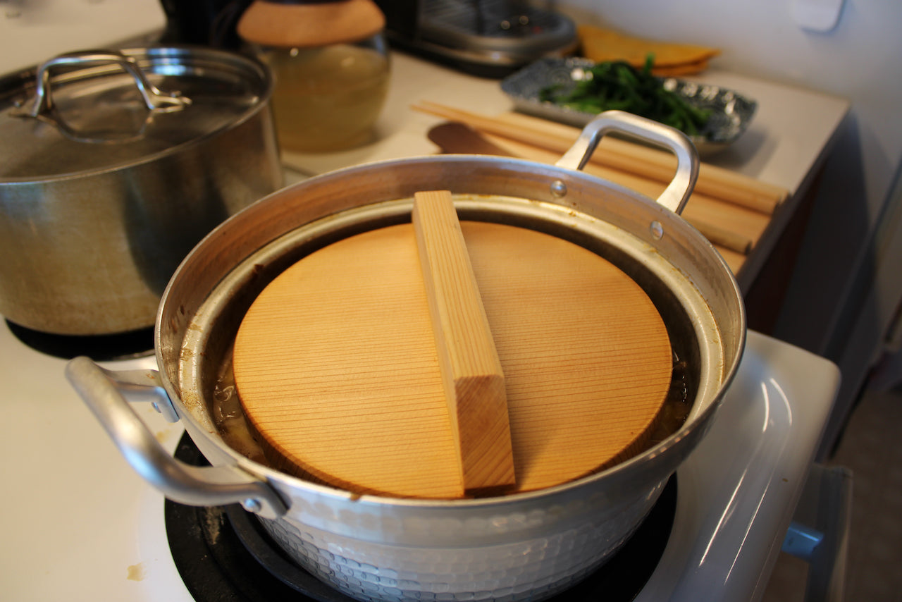 round wooden drop inside two handled pot on stovetop surrounding cookware vegetables and utensils in kitchen background