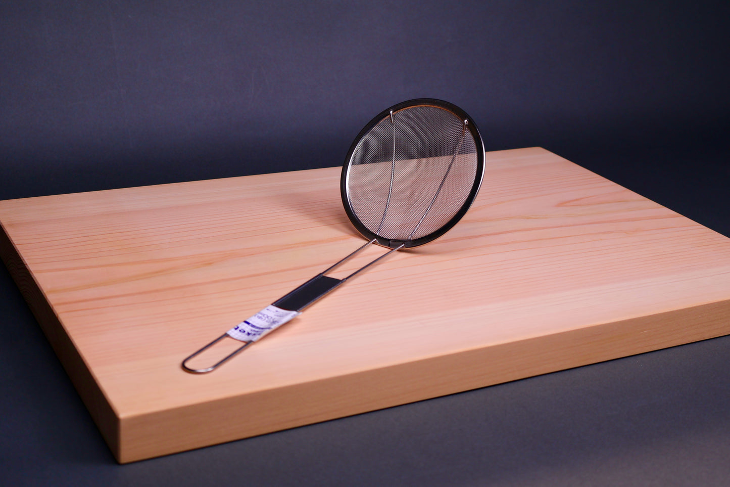 small fine meshed stainless steel skimmer sitting atop hinoki cutting board in grayish blue background