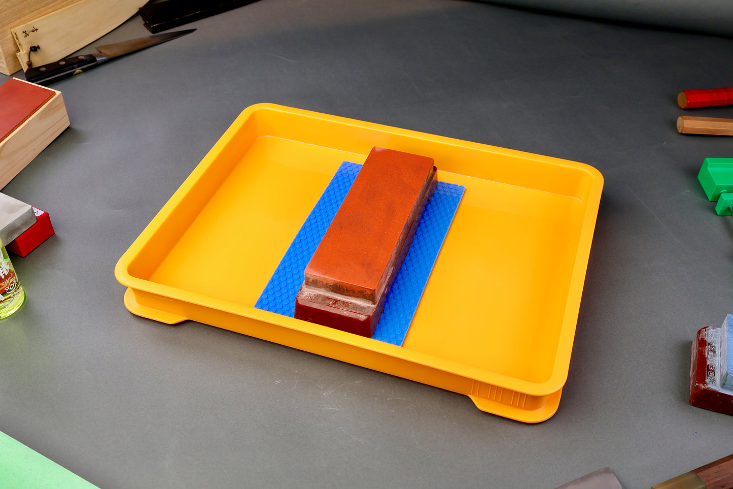shot and wide yellow container holding hasegawa non slip mat and naniwa whetstone surrounded by knife accessories and grey backdrop