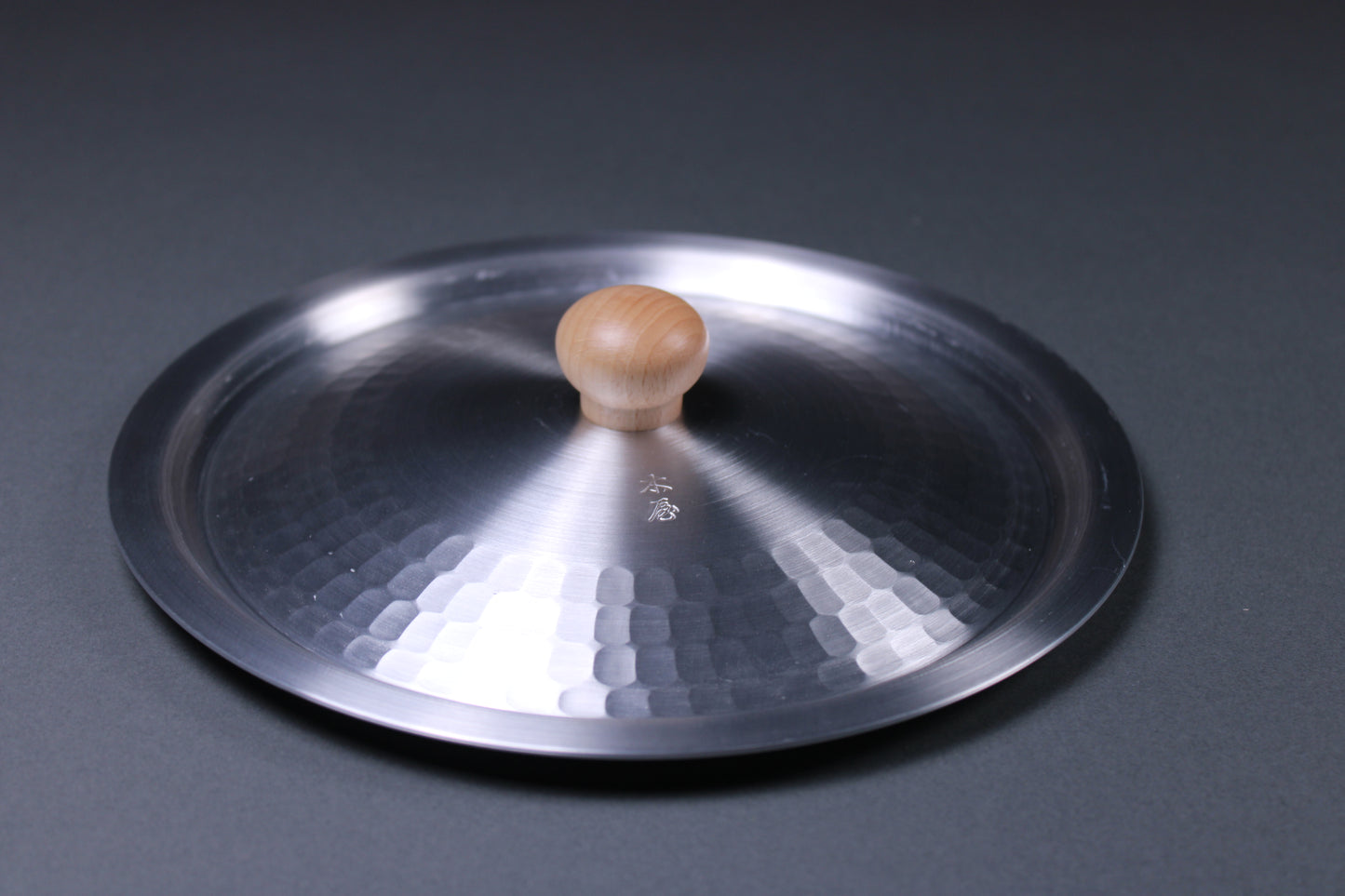 round metallic lid with hammered interior portion with wood knob in center sits on gray surface