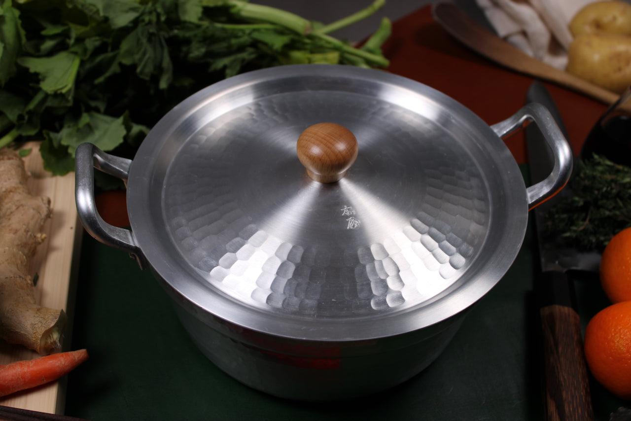round metallic hammered lid with wood knob in center sits atop two handled metal pot surrounded by produce in dark setting