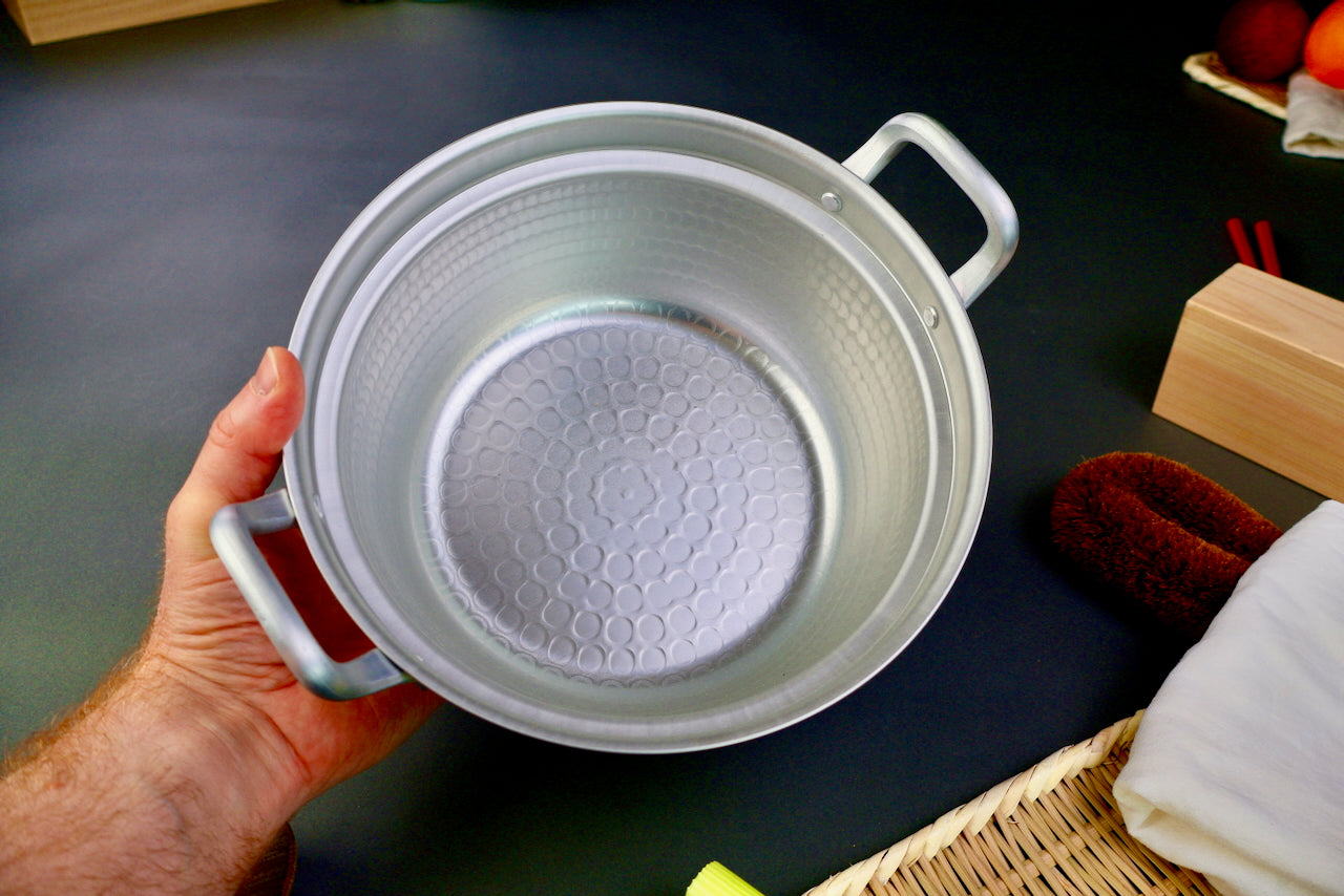 hand holding two handled stepped aluminum silvery pot with shiny hammered surface dark grey backdrop