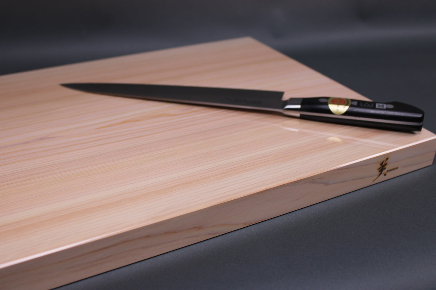 rectangular shaped light medium deep wood tan hinoki cutting board with deep natural grain lines black handled knife atop board in front of dark grey backdrop