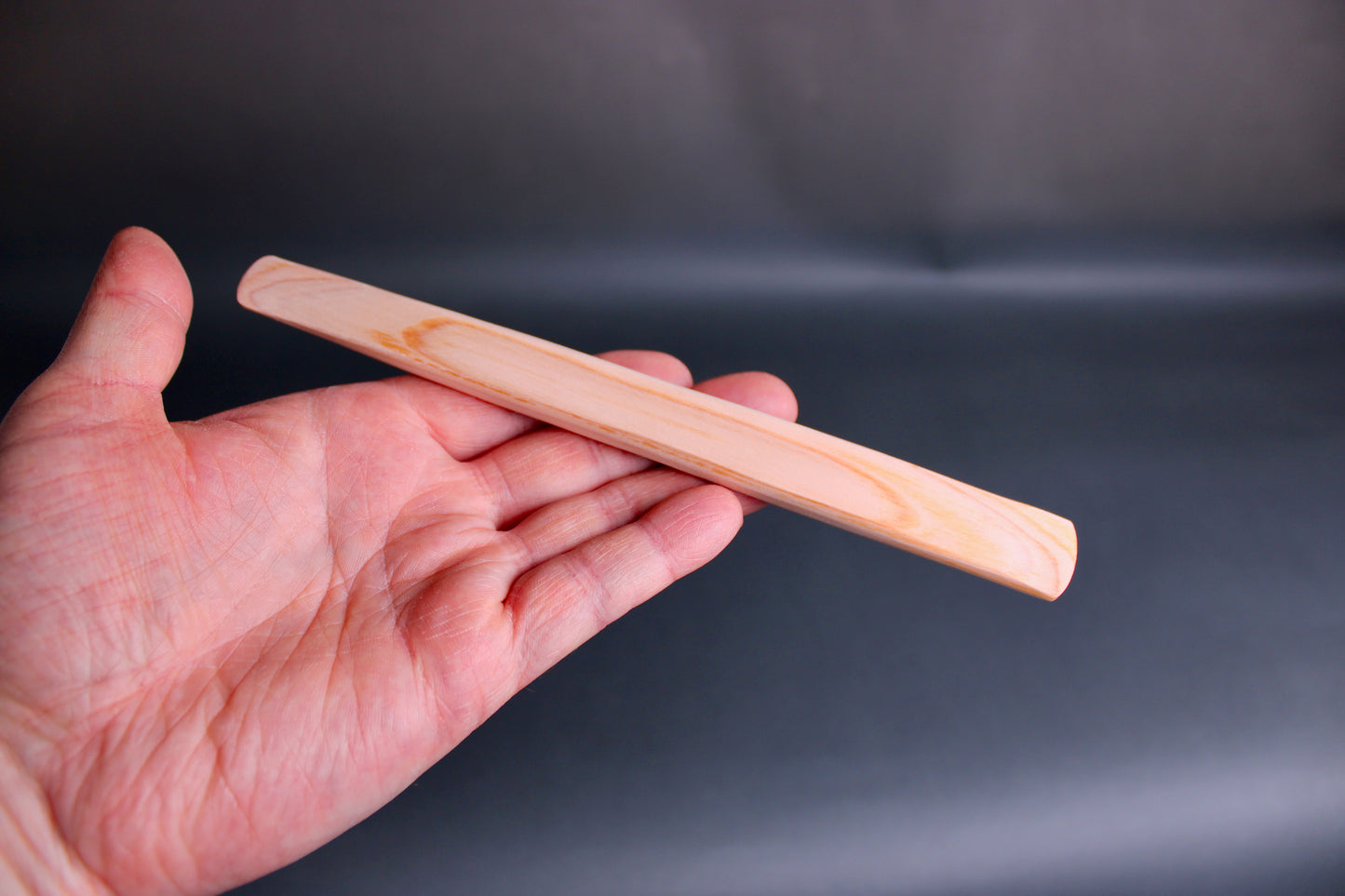 hand holding long thin flat natural wood tone hinoki spreader in dark grey backdrop