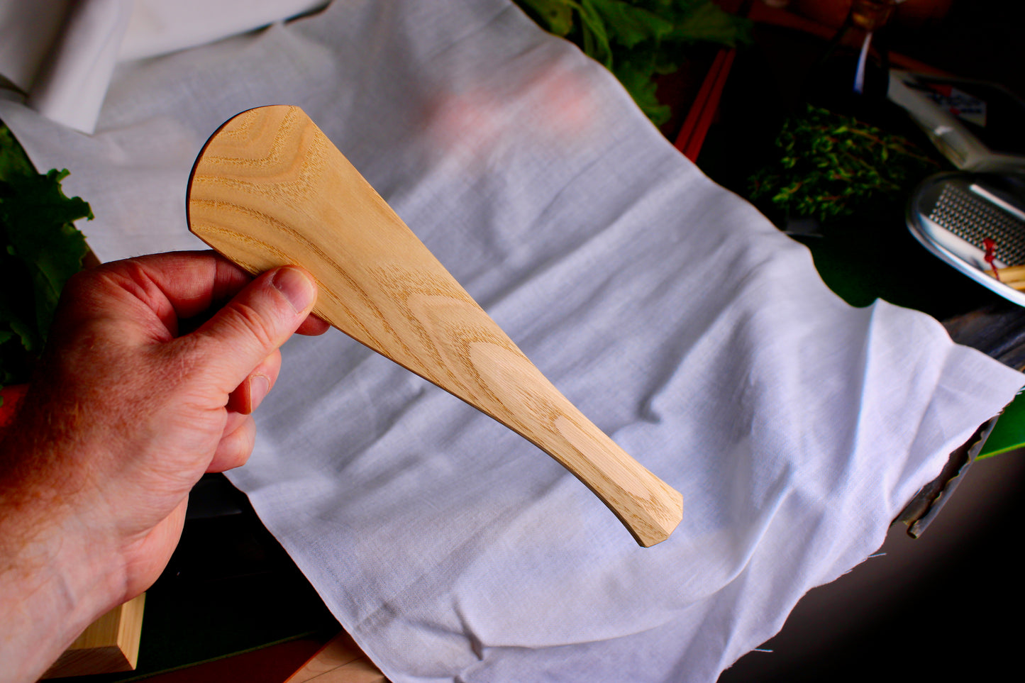 hand holding a long wooden tan rice scoop spoon showing wood grain on white cloth background with kitchenware