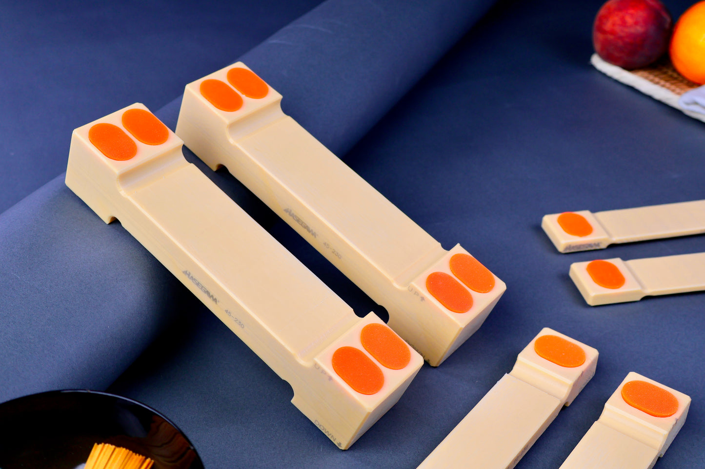 three sizes of hasegawa cutting board lifters long rectangular shaped with pill shaped bright orange oval non slip material in blue grey background with fruit and towel