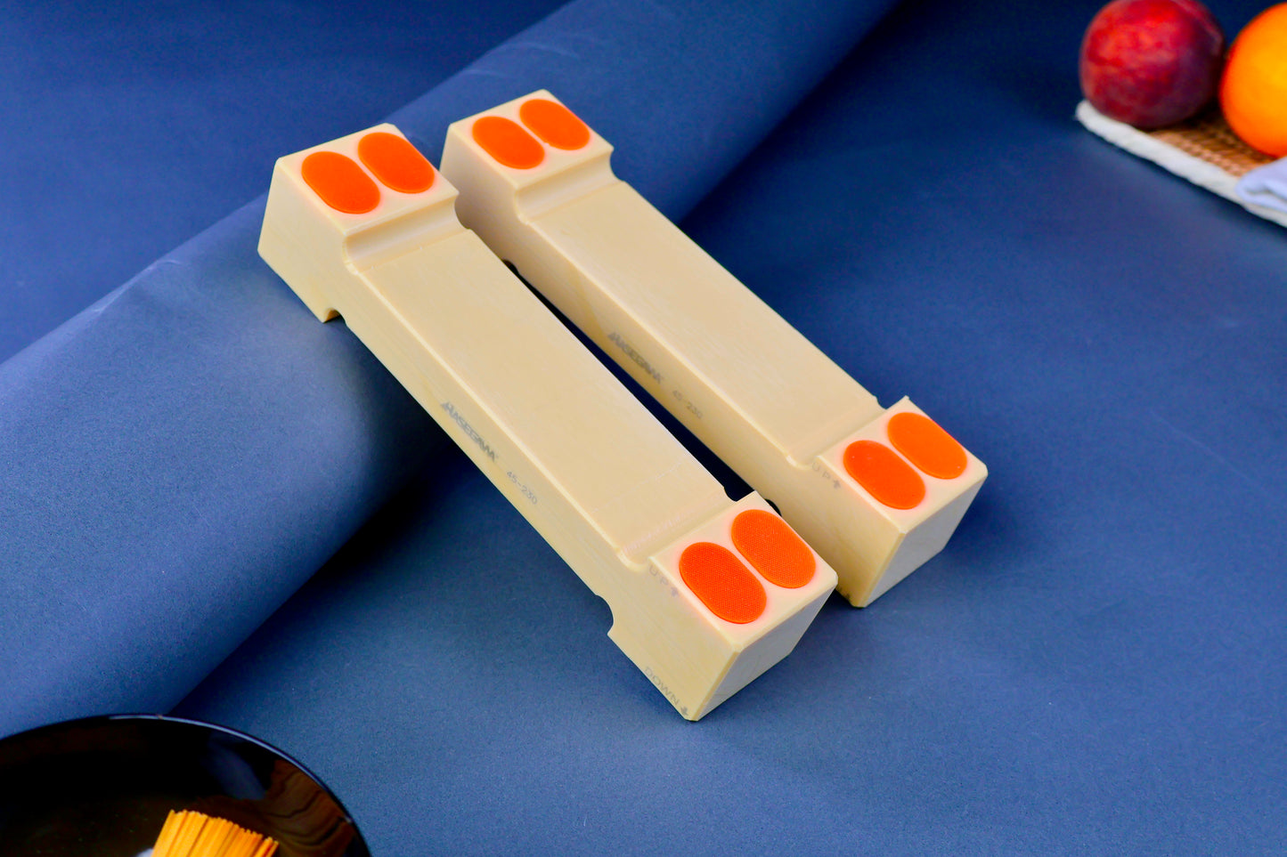 hasegawa cutting board lifter forty five mm tall long rectangular shaped with pill shaped bright orange oval non slip material in blue grey background with fruit and towel