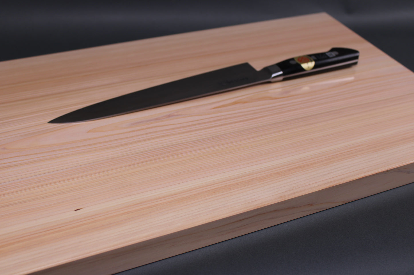 rectangular shaped light medium deep wood tan hinoki cutting board with deep natural grain lines knife sitting atop in front of dark grey backdrop