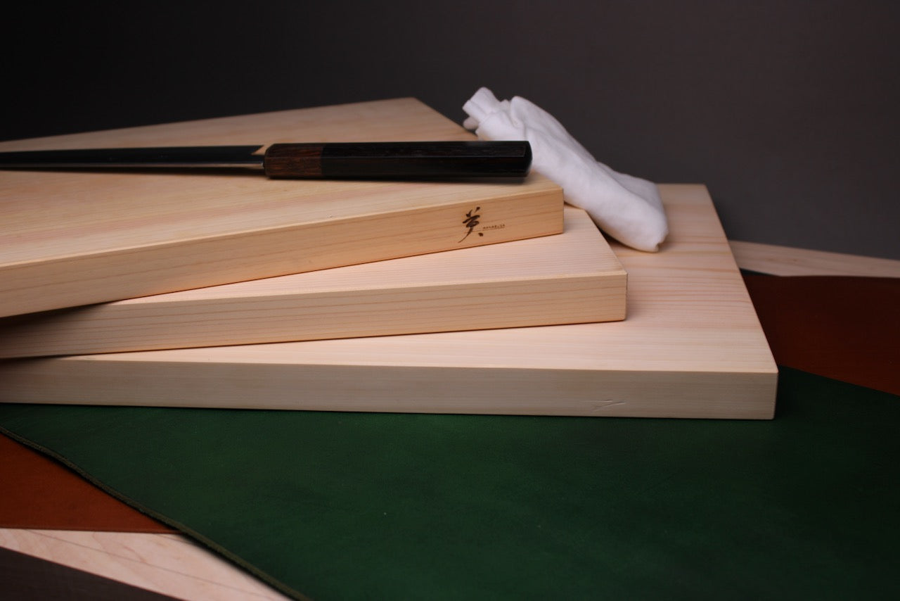 three rectangular shaped light medium deep wood tan hinoki cutting boards dark knife atop white towel behind in front of dark grey backdrop