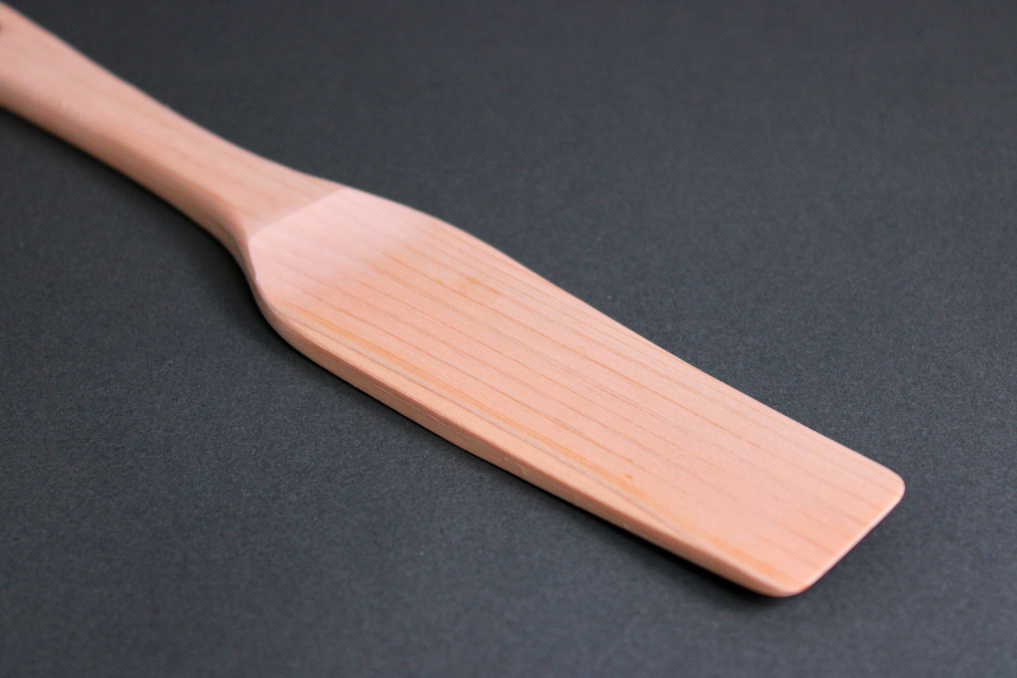 close up of small hinoki wood spatula long thin handle and slightly larger broad spatula area on dark grey surface