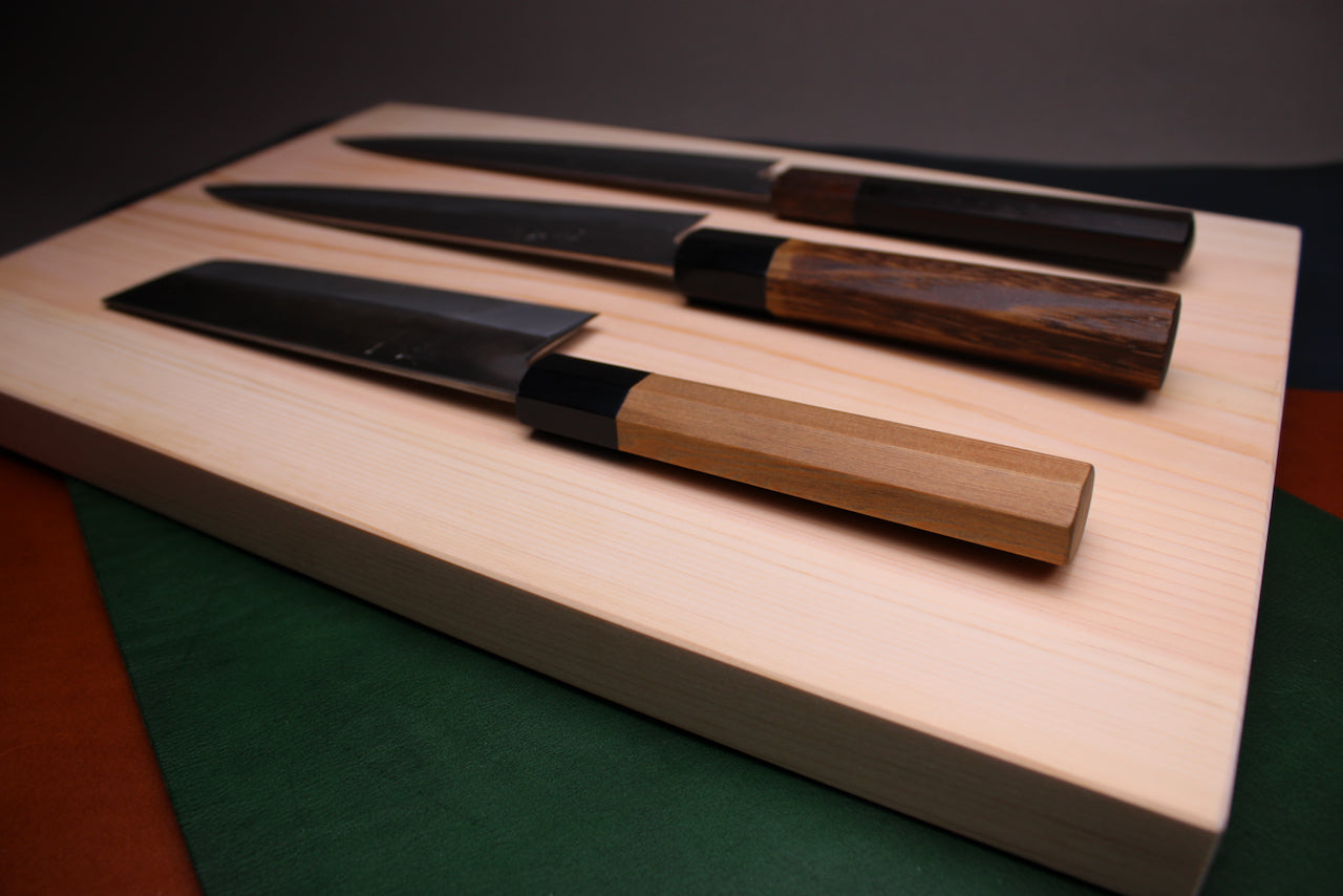 three wood handled knives atop rectangular shaped light medium deep wood tan hinoki cutting board with deep natural grain lines in front of dark grey backdrop