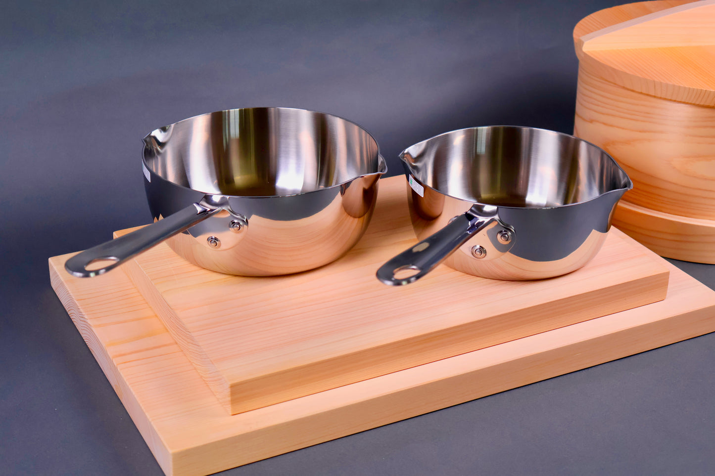 two steel shiny long handled saucepans on hinoki cutting boards beside seiro steamer with grey background