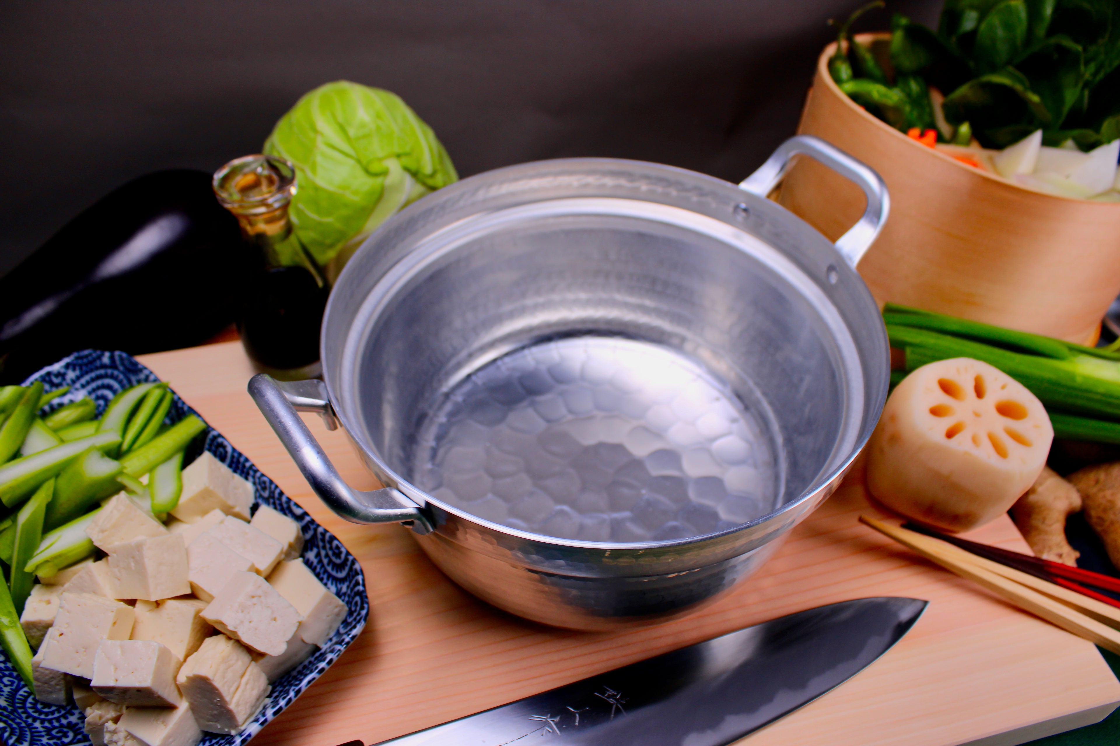 shiny bright two handled aluminum hammered pot atop tan wood cutting board surrounding various ingredients tofu cabbage with knife chopsticks in foreground