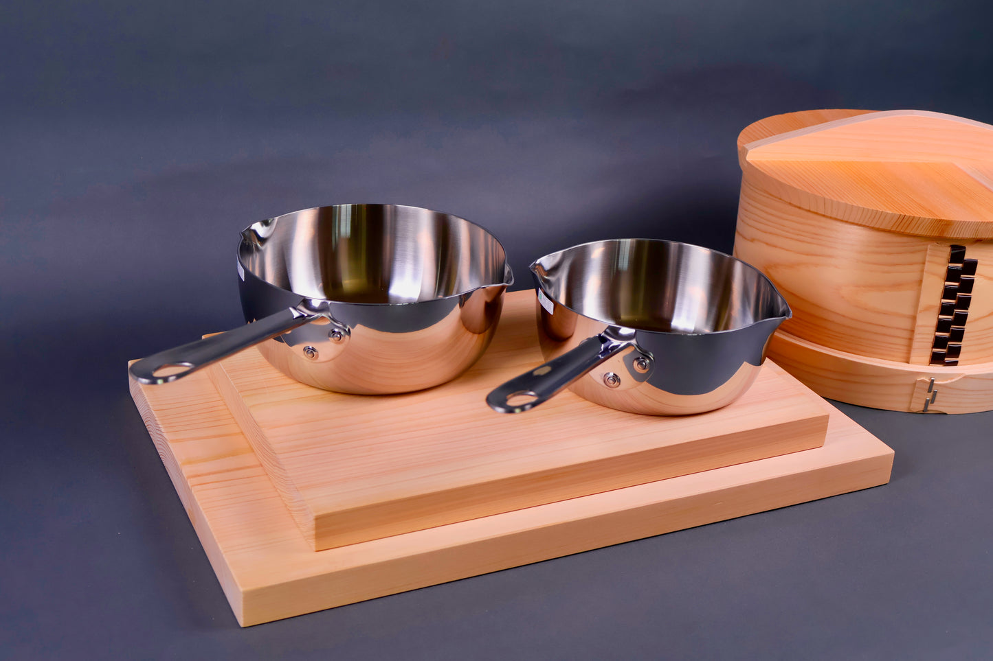 two japanese cooking saucepans shiny silver on cutting boards beside wood container in grey background