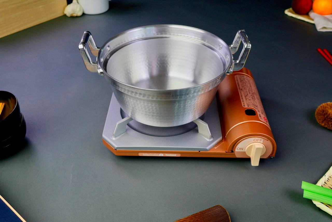 two handled stepped aluminum silvery pot with shiny hammered surface sits atop portable silver tan butane stove on a dark grey backdrop