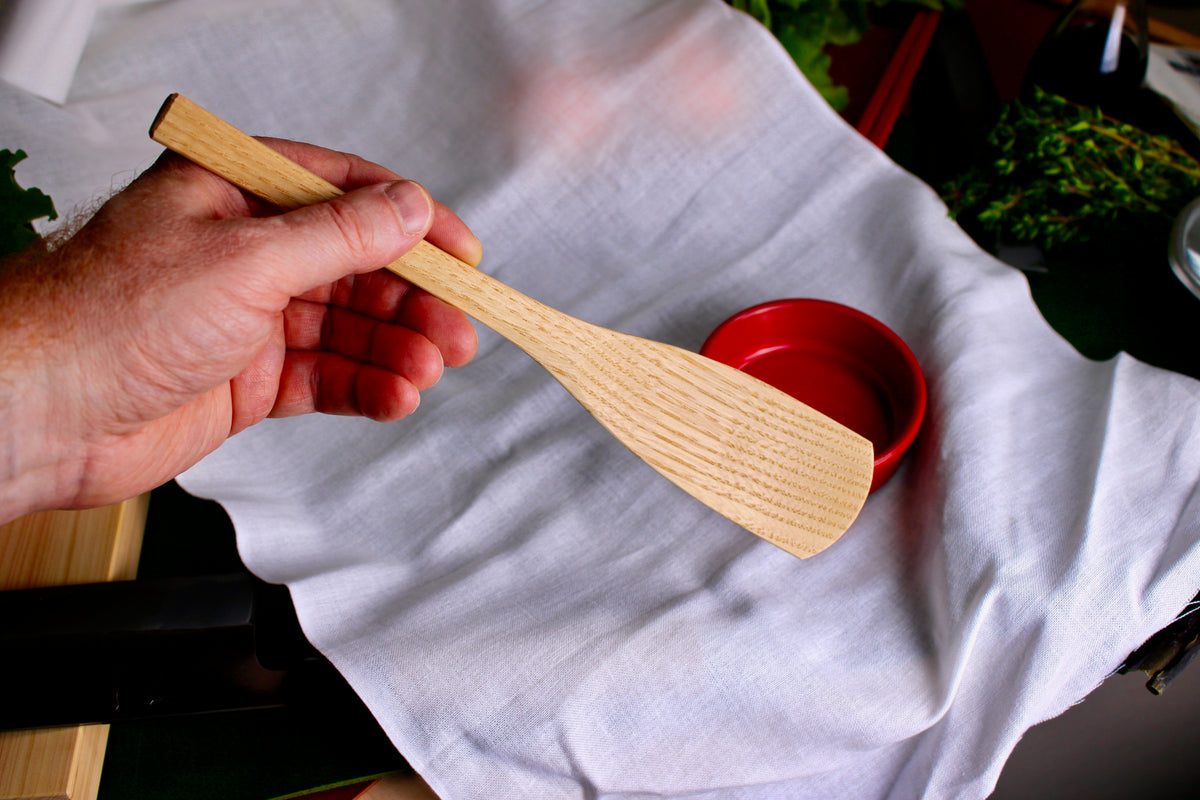hand holding a long smooth planed wooden spoon showing detailed wood grain pattern broad thin end for scoop contrasted on white cloth backdrop