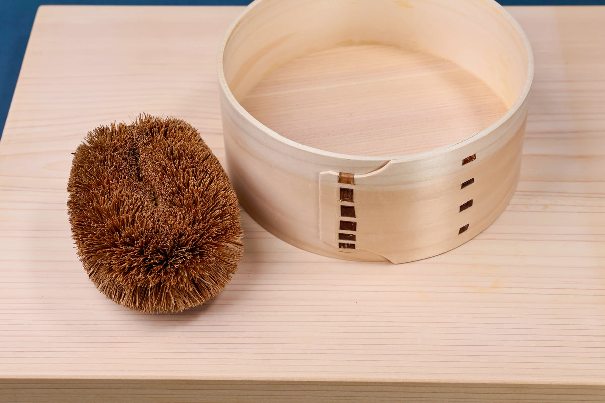 Kamenoko Tawashi scrubber resting beside cedar seiro steamer body on hinoki cutting board with blue background
