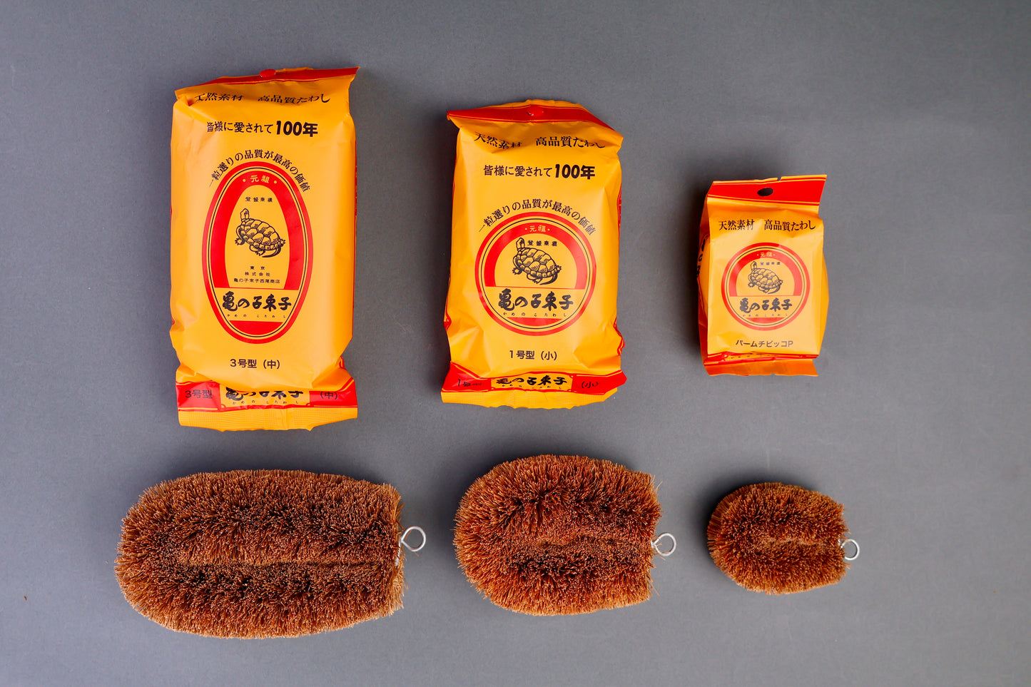 three yellow red plastic packages beside tawashi palm brushes each package with an animated image of a turtle with kanji characters on grey background