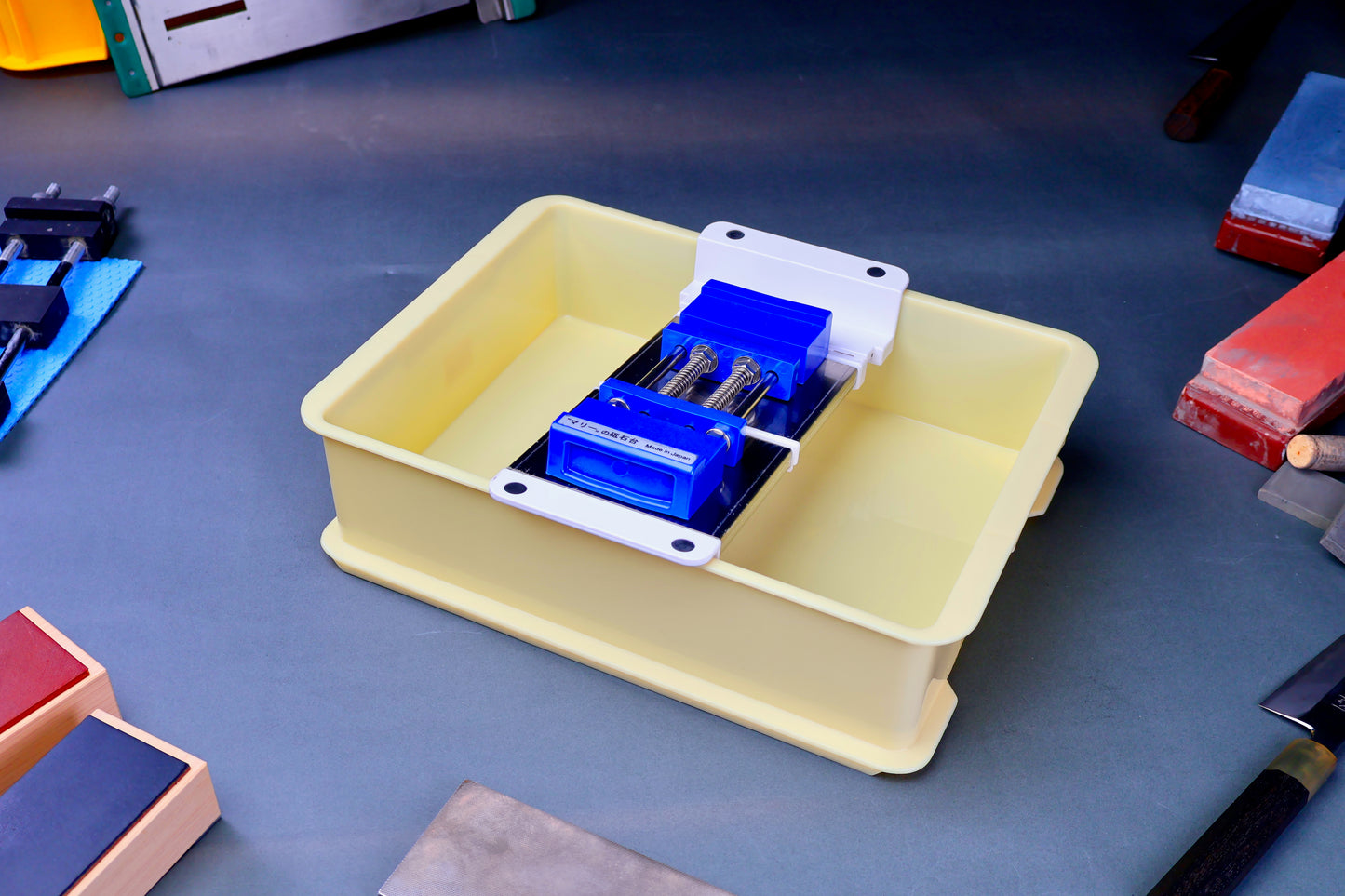 short yellow plastic container with suehiro multi sliding rack and blue rubber whetstone holder surrounded by knives and accessories on blue grey surface