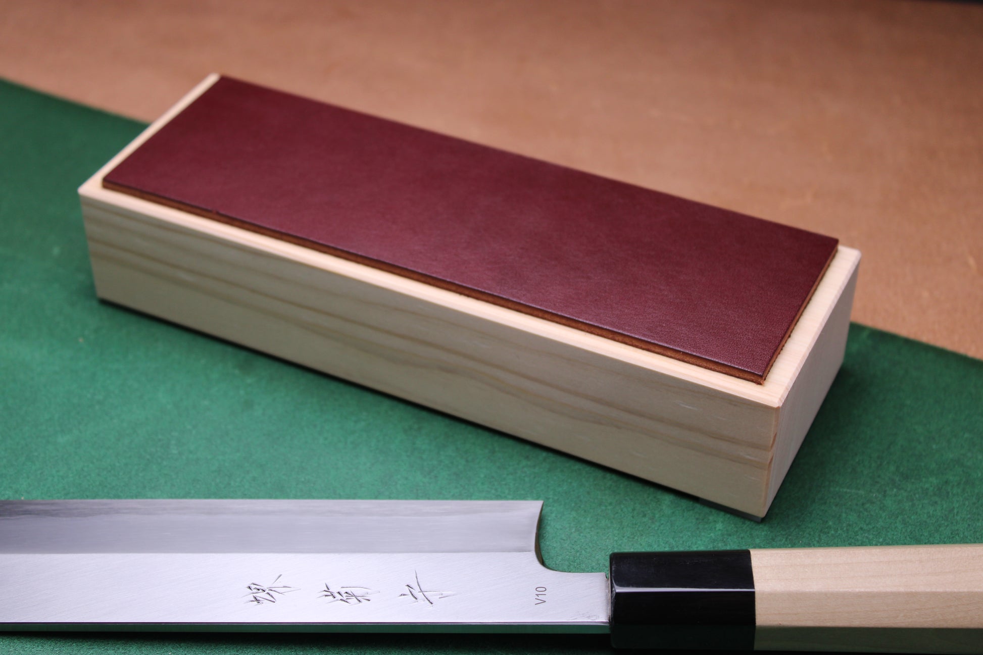 knife strop made of English bridle leather colored burgundy espresso brown attached to sturdy tall smooth hinoki wood base sakai kikumori japanese usuba vegetable knife foreground with background faded green and brown