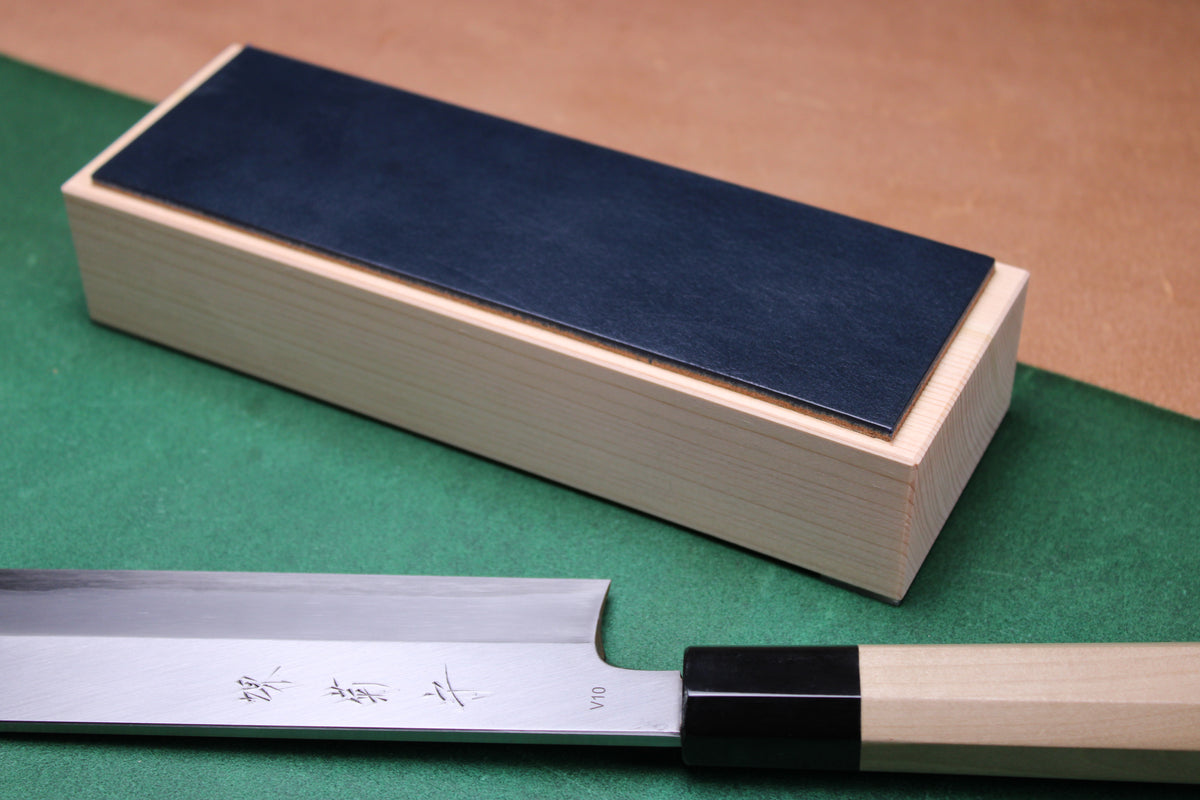 knife strop made of English bridle leather colored midnight blue attached to sturdy tall smooth hinoki wood base sakai kikumori japanese usuba vegetable knife foreground with background faded green and brown