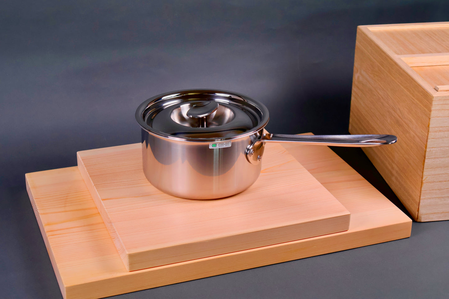 lidded saucepan shiny exterior art deco style atop boards beside wood box grey background