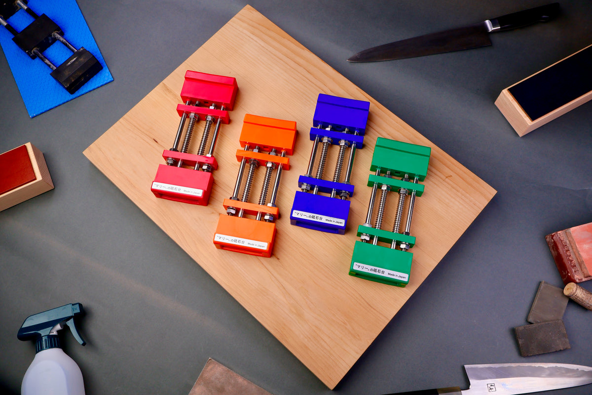 red orange blue and green sharpening stone holder atop hinoki cutting board surrounded by knife sharpening accessories and grey background
