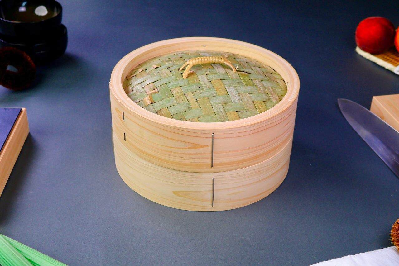 thick wood round bamboo steamer set woven top with loop natural wood tone with printed hiragana surrounded by japanese kitchenware blue gray background