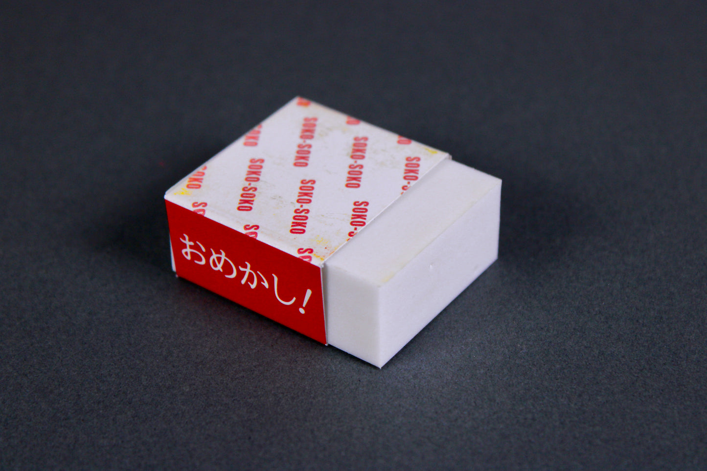 short rectangular white rust eraser partial wrapped product paper printed soko soko hiragana omekashi on dark grey background