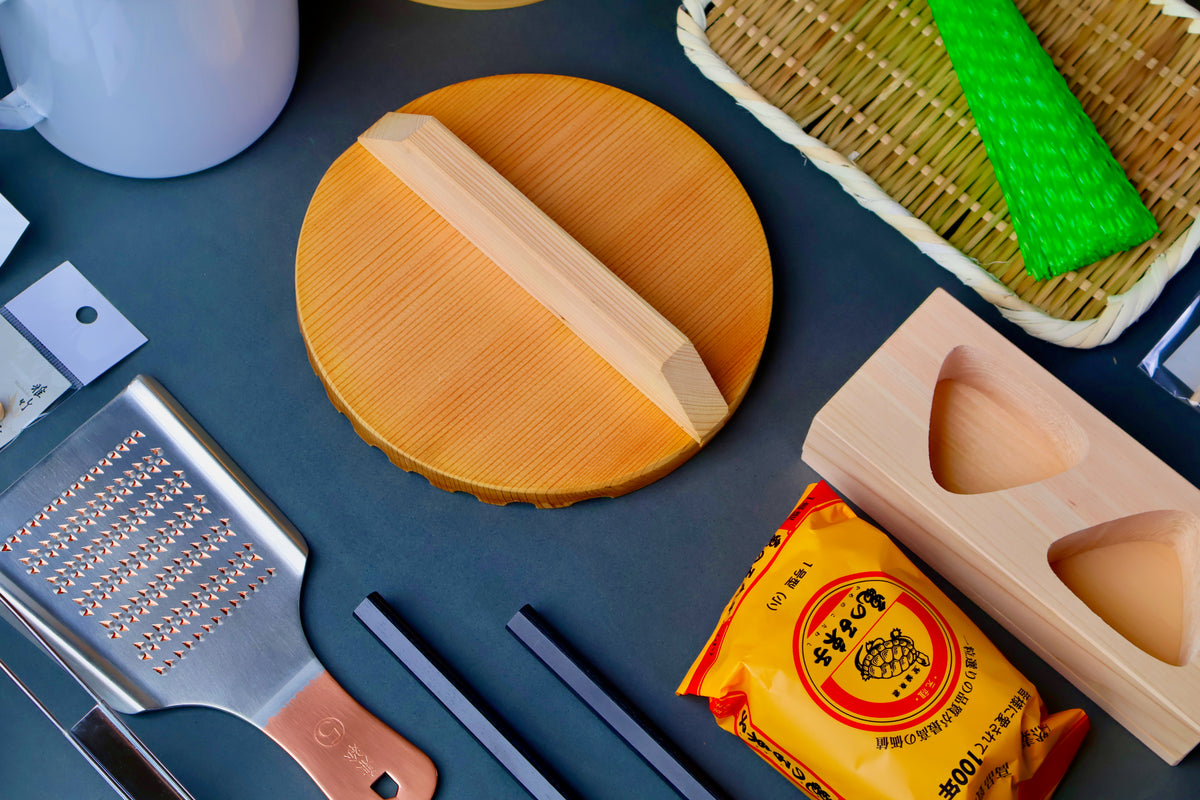 circular wood drop lid with handle copper metal square good grater bamboo strainer with bright green brush alongside other kitchenware on dark backdrop