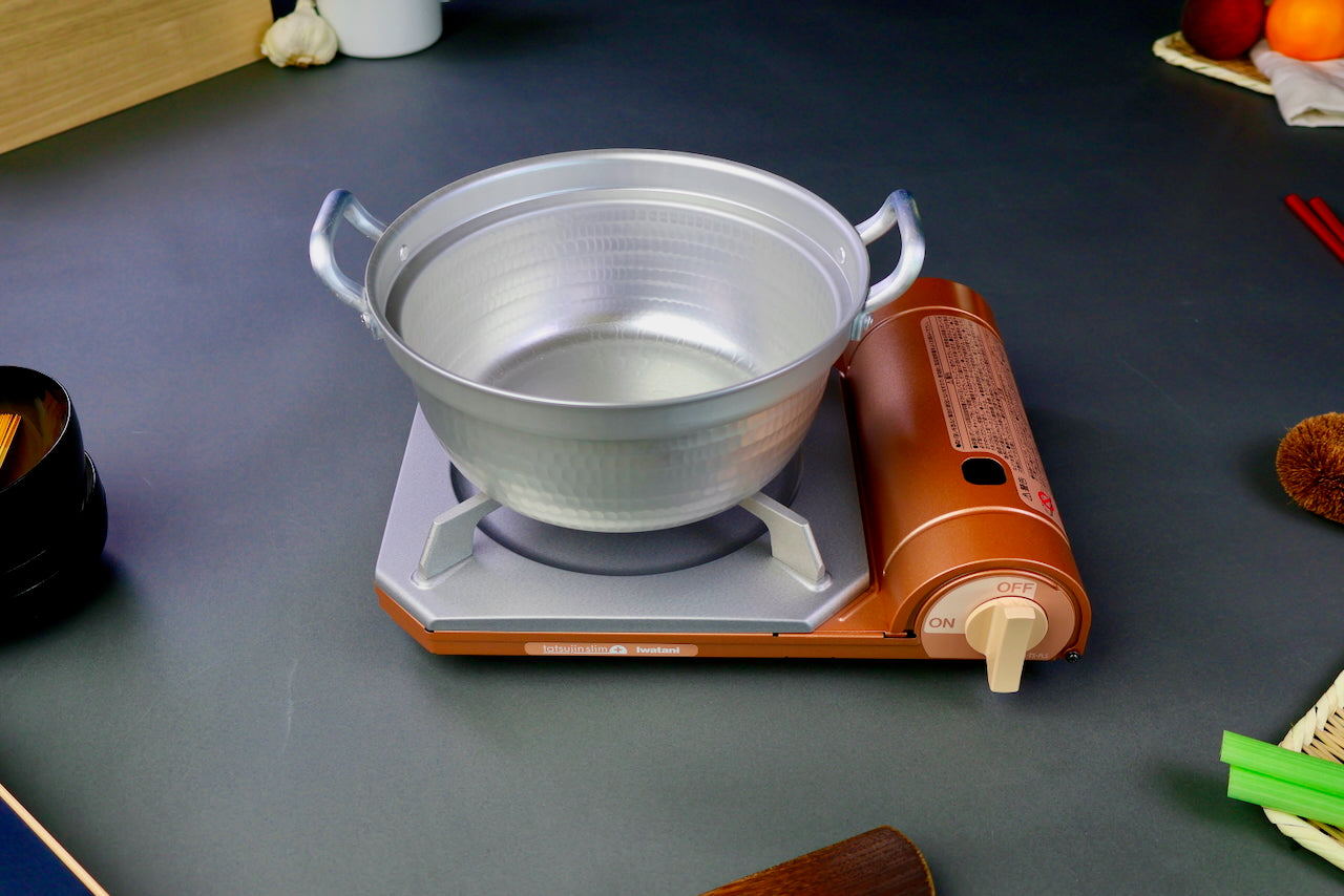 two handled stepped aluminum silvery pot with shiny hammered surface sits atop portable silver tan butane stove on a dark grey backdrop