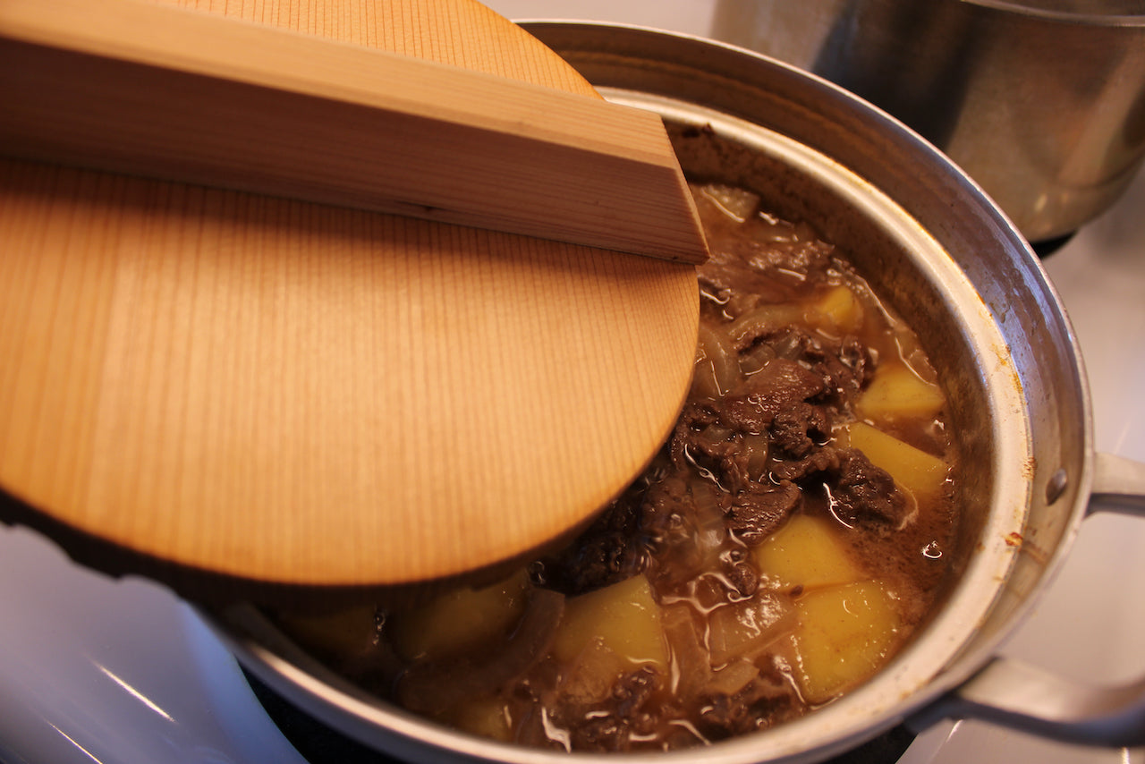 round wooden drop being removed from two handled pot on stovetop showing contents of beef and potatoes in kitchen background