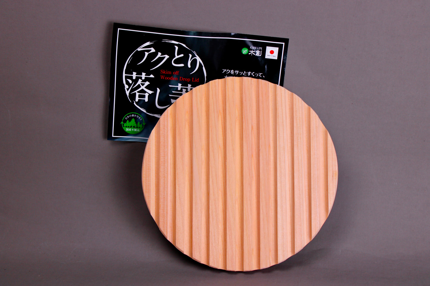 back side with ridges circular wood drop lid with long shallow handle yellowish gold hue beside black product tag with hiragana in front of grey background