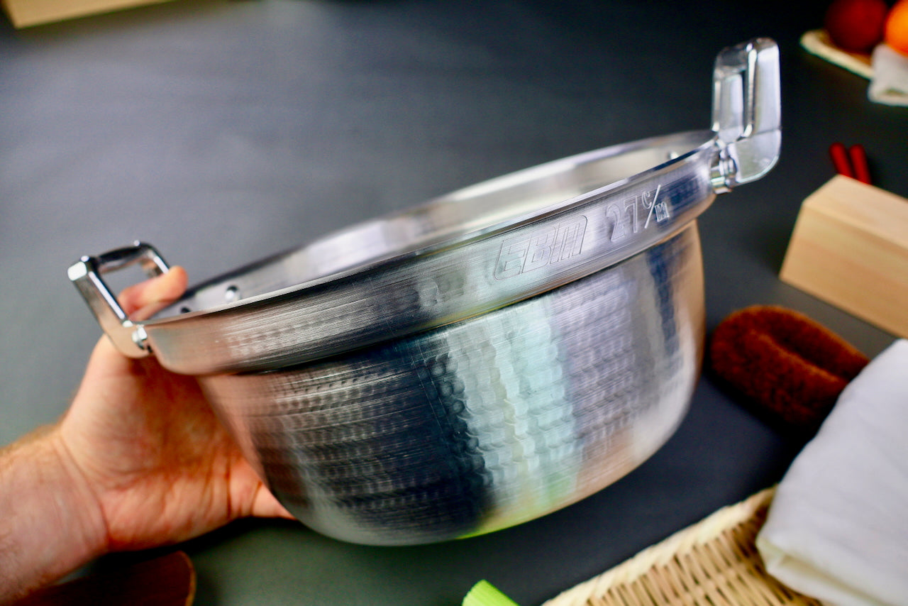close up of hand holding stepped aluminum silvery pot with shiny hammered surface on a dark grey backdrop