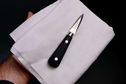 hand holding white towel on which sits black handled steel oyster knife engraved in French with beveled edges dark background