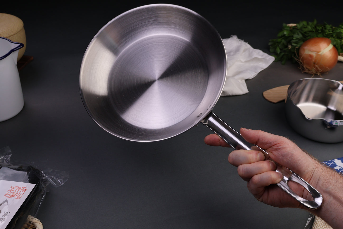 Torino tag brand saute pan held in hand centered around yukihira saucepan vegetables pitcher in dark background